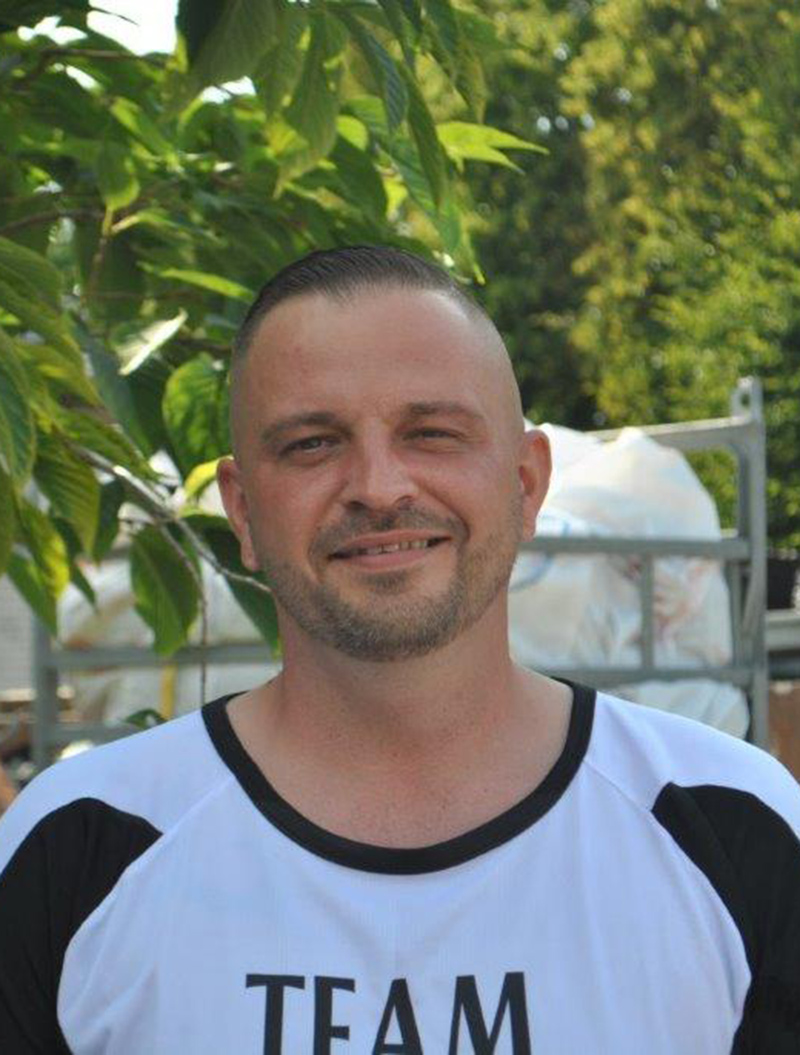 Mann lächelt leicht, trägt schwarz-weißes Shirt, kurze Haare, draußen vor grünen Blättern.