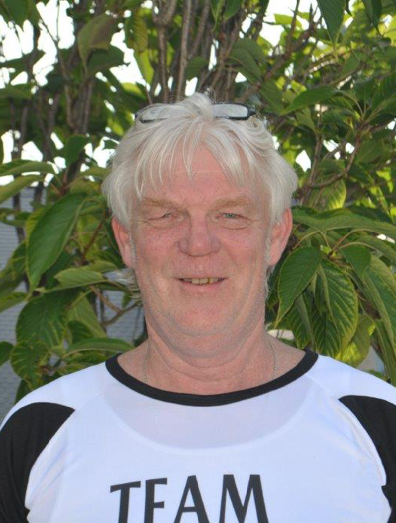Älterer Mann lächelt vor grünen Blättern; trägt schwarz-weißes Shirt und Brille auf Kopf.