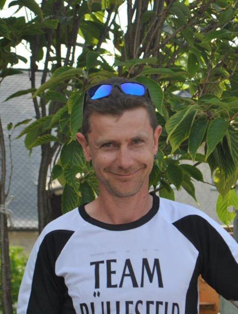 Ein lächelnder Mann mit Sonnenbrille auf dem Kopf, in einem schwarz-weißen Shirt, vor Blättern.