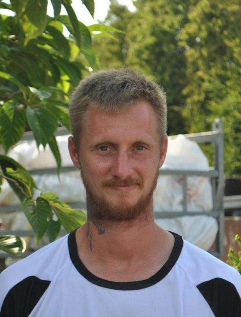 Ein lächelnder Mann mit kurzem Haar und Bart, trägt ein weißes T-Shirt, steht vor grünen Blättern.