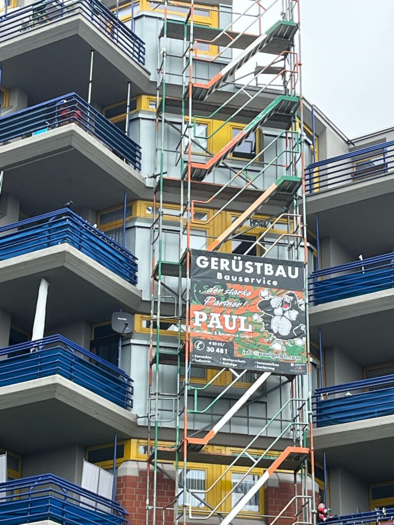 Ein Baugerüst mit bunten Balkonen eines Gebäudes, Werbeschild von Gerüstbauservice.