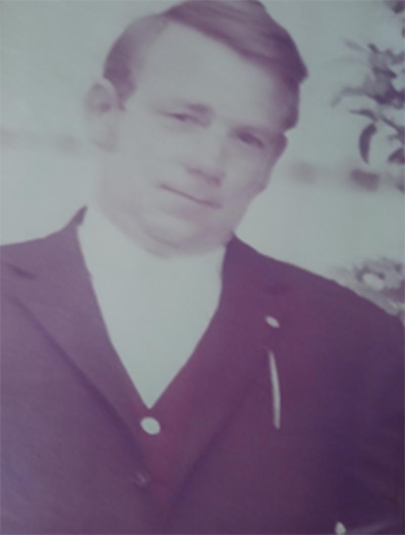 Ein Mann in Anzug mit neutralem Ausdruck, lila Farbton, unscharf hinten, Blumen rechts.