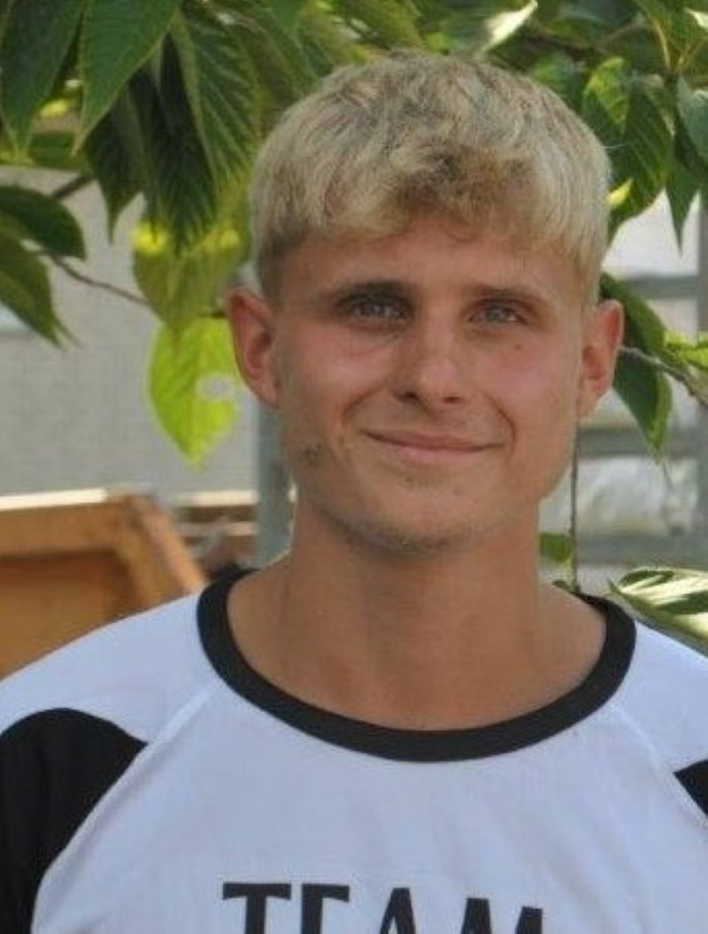 Laechelnder blonder junger Mann im weissschwarzen Trikot vor gruenem Laub
